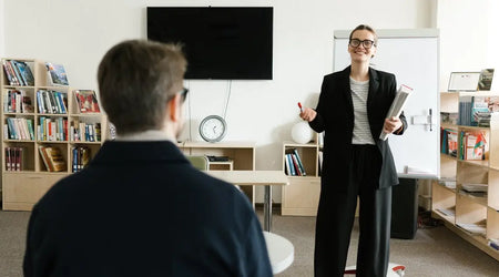 Smiling woman teaching man in classroom with bookshelves and whiteboard in background